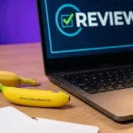 “Gemini 3 Nano Banana Pro placed next to a real banana, a notebook, and a laptop displaying ‘REVIEW’ on the screen on a wooden desk.”